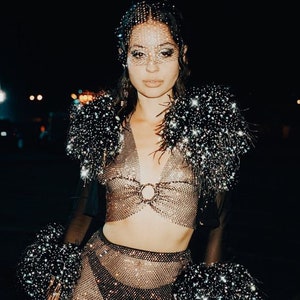 May include: A woman wearing a sparkly, sheer, mesh top and skirt with a black feather trim. She is wearing a crystal-encrusted face mask.