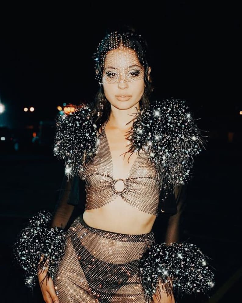 May include: A woman wearing a sparkly, sheer, mesh top and skirt with a black feather trim. She is wearing a crystal-encrusted face mask.