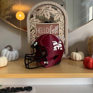May include: A burgundy football helmet with a black facemask and white logo. The helmet is displayed on a wooden shelf with decorative pumpkins and a wooden sign that says "Hello Fall".