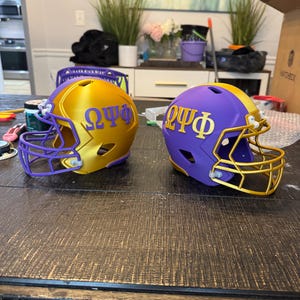 May include: Two miniature American football helmets, one gold and one purple, are displayed on a dark wooden surface. Each helmet has a face mask and the Greek letters ΩΨΦ. The gold helmet has purple accents, while the purple helmet has gold accents.