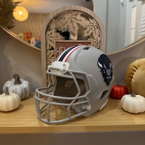 May include: A gray football helmet with a navy blue buffalo logo and red and white stripes. The helmet is displayed on a wooden shelf with decorative pumpkins and a mirror in the background.