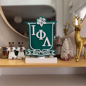 Könnte beinhalten: Ein grünes und weißes Sorority-Wappen mit einer weißen Rose und den Buchstaben "IΦΛ" sowie dem Text "EST. 1929".