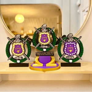 Peut inclure: Trois trophées dorés et violets avec un bouclier, des épées et une couronne. Le bouclier porte les lettres "ΩΨΦ" et une lampe. Le texte "Chapter or Line Name Here Omega Psi Phi Fraternity, Inc" est inscrit sur la base du trophée central.