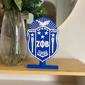 May include: A blue and white decorative shield with Greek text, stars, and the letters ZΦB, and the year 1920. The shield is mounted on a blue base and sits on a wooden shelf. A clear glass vase with white flowers is in the background.