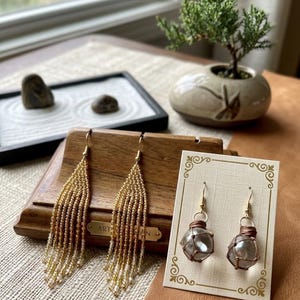 May include: Two pairs of earrings: one pair features long, gold-toned beaded fringe, and the other pair consists of clear glass spheres wrapped in brown cord, displayed on a white card with gold decorative borders and gold-toned metal hooks.