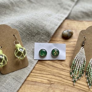 May include: Three pairs of earrings: one pair of clear glass spheres wrapped in light green cord, one pair of round green studs with gold flecks, and one pair of long, white and green ombre beaded fringe earrings on brown cardstock.