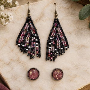 May include: A pair of long, triangular fringe earrings crafted from black, pink, and white seed beads, paired with a set of round stud earrings featuring a textured, shimmering mauve-pink surface with gold-toned flecks and dark metal settings.