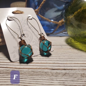 May include: Teal glass beads wrapped in brown twine, hanging from silver earring hooks. The earrings are displayed on a white card with the letter "F" printed on it.