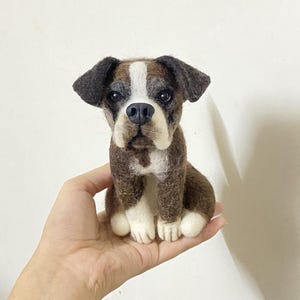 May include: A brown and white needle felted boxer dog figurine. The dog is sitting with its paws crossed and has a white patch on its chest.