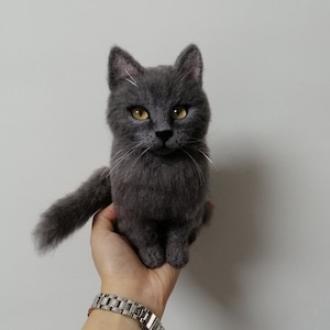 Könnte beinhalten: Eine graue, gefilzte Katzenfigur mit gelben Augen. Die Katze sitzt auf der Hand einer Person.