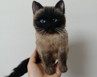 May include: A hand-crafted needle felted Siamese cat with blue eyes. The cat is sitting up with its tail curled around the hand holding it.