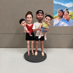 May include: A family figurine featuring two adults and two children, all with smiling faces. The adults are wearing casual clothing, and the children are being held. The figurine is on a round, gray base, with a photo of the family in the background.
