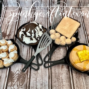 Puede incluir: Una variedad de desayunos en miniatura en sartenes de hierro fundido negro, que incluyen rollos de canela, postre de chocolate con crema batida, tostadas con huevos y panqueques con mantequilla. Cada sartén tiene una espátula.