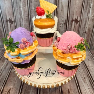 Könnte beinhalten: Drei dekorative Cupcakes auf einem weißen und goldenen Kuchenständer. Die Cupcakes haben Waffelböden, rosa und lila Zuckerguss und sind mit Beeren, Grünzeug und einer Waffel belegt. Die Cupcakes stehen auf einer Holzoberfläche.