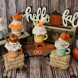 May include: Five decorated mugs with autumn-themed designs and toppings, including pumpkin spice, candy corn, and cinnamon sticks. The mugs are arranged on a wooden surface with a wooden sign that reads "hello fall".