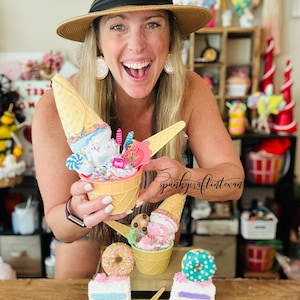 Könnte beinhalten: Eine Frau hält mehrere dekorative Eiscreme- und Kuchenartikel. Die Artikel umfassen gefälschte Eiswaffeln, Kuchenstücke und einen Donut, alle in Pastellfarben mit Streuseln und dekorativen Elementen. Die Artikel werden auf einer Holzoberfläche präsentiert.
