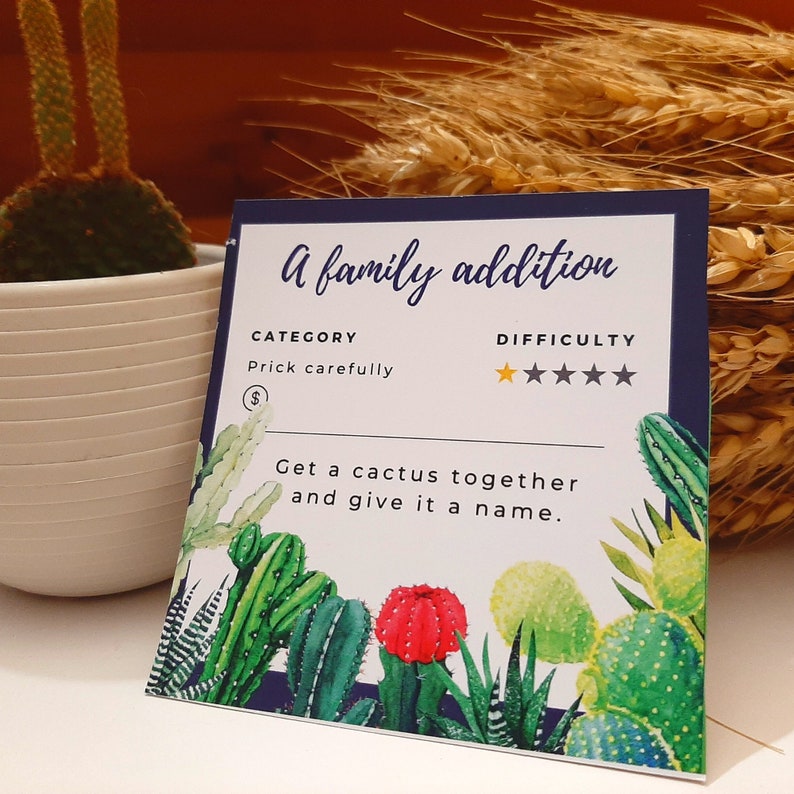 Op de afbeelding: Een witte kaart met een blauwe rand en een cactus-illustratie. De kaart luidt "A family addition", "Categorie: Prick carefully", "Moeilijkheid: 5 sterren", en "Get a cactus together and give it a name."