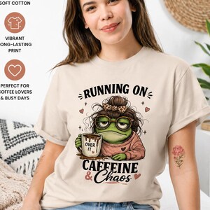 Pode incluir: Camiseta bege com o texto "RUNNING ON CAFFEINE & Chaos". O gráfico apresenta um sapo usando óculos e segurando uma caneca de café. A camisa também tem ícones indicando algodão macio premium e impressão vibrante e duradoura.