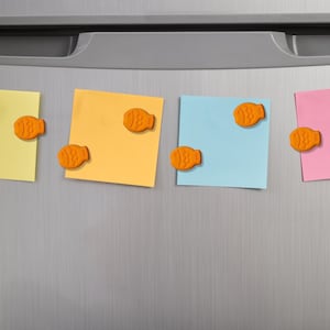 Op de afbeelding: Vier gekleurde plaknotities, geel, oranje, blauw en roze, zijn bevestigd op een zilveren koelkastdeur. Elke notitie wordt op zijn plaats gehouden door een oranje, visvormige magneet. De notities zijn gelijkmatig verdeeld.
