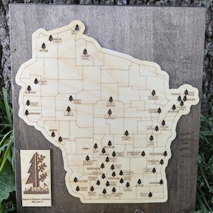 May include: Wooden map of Wisconsin with engraved county lines and tree icons. The map is mounted on a dark stained wooden board. A smaller wooden plaque with a tree and text is attached. The text on the plaque reads "Karen & Stephen Swatlen ABJ part II."