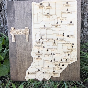 May include: A wooden map of Indiana with the names of various lakes and towns throughout the state. The map is mounted on a brown wooden background. The text "Our Adventures" is written on a small wooden sign.