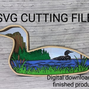 May include: SVG cutting file of a loon on a lake with a forest and mountains in the background. The loon is black and white with a red eye. The lake is blue and the forest is green. The mountains are brown. The text "SVG CUTTING FILE" is at the top of the image. The text "Digital download not finished product" is at the bottom of the image.