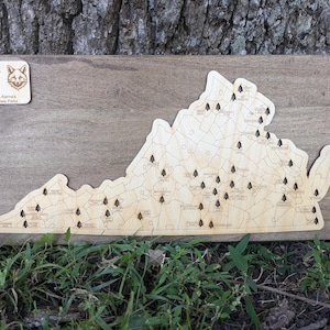 May include: A wooden map of Virginia with small pine tree icons marking the locations of dog-friendly state parks. The map is mounted on a wooden board. A small wooden sign with the text "Paw & Paws VA State Parks" and illustrations of a golden retriever and a fox is in the top left corner.