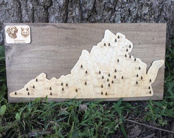 May include: A wooden map of Virginia with small pine tree icons marking the locations of dog-friendly state parks. The map is mounted on a wooden board. A small wooden sign with the text "Paw & Paws VA State Parks" and illustrations of a golden retriever and a fox is in the top left corner.