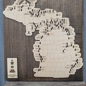 May include: Wooden map of Michigan with lighthouse markers. The map is mounted on a dark wooden background. A small wooden plaque with a lighthouse illustration and the text "The Brouwer's Michigan Lighthouse Travels" is on the left.