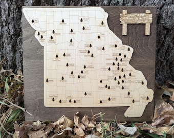 May include: A wooden map of Missouri with the names of state parks marked with small trees. The map is mounted on a brown wooden background with a sign that reads "Miles & Family State Park Adventures".
