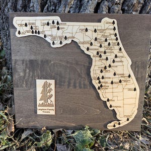 May include: A wooden map of Florida with small black trees marking locations. The map is mounted on a dark brown wooden plaque. A smaller plaque below the map reads "Leighton Family Travels" with a small tree graphic.