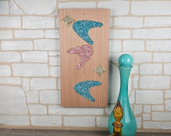 May include: A wooden wall art piece with three boomerang shapes made of colorful mosaic tiles. The boomerangs are in shades of blue, pink, and white. The piece is mounted on a light brown wooden background.