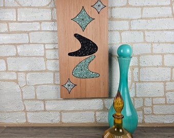 May include: A wooden wall plaque with a mid-century modern design featuring two boomerang shapes made of turquoise and black mosaic tiles. The plaque is decorated with four star shapes made of turquoise mosaic tiles. The plaque is hanging on a white brick wall.