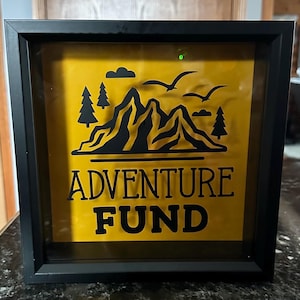 Op de afbeelding: Zwarte schaduwbox met gele achtergrond. De box heeft een zwarte bergketen, bomen, vogels en de woorden "ADVENTURE FUND" in een vetgedrukte lettertype. Een decoratief stuk om geld te sparen voor reizen.