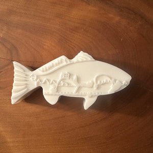 May include: A white, fish-shaped decorative object with detailed relief carvings. The design includes a depiction of a landscape within the fish's body. The object is set against a warm, wooden background.