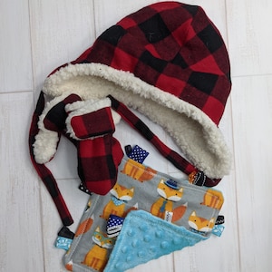 May include: A red and black plaid hooded hat with a white sherpa lining, matching mittens, and a fox-themed baby blanket with a blue minky side.