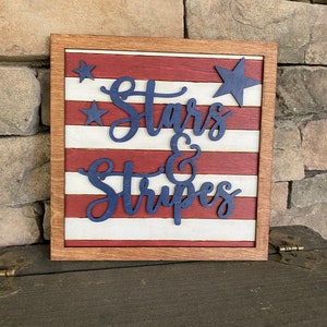 Puede incluir: Un letrero de madera con un dise&ntilde;o de bandera estadounidense roja, blanca y azul. El letrero dice "Stars & Stripes" en letras azules.