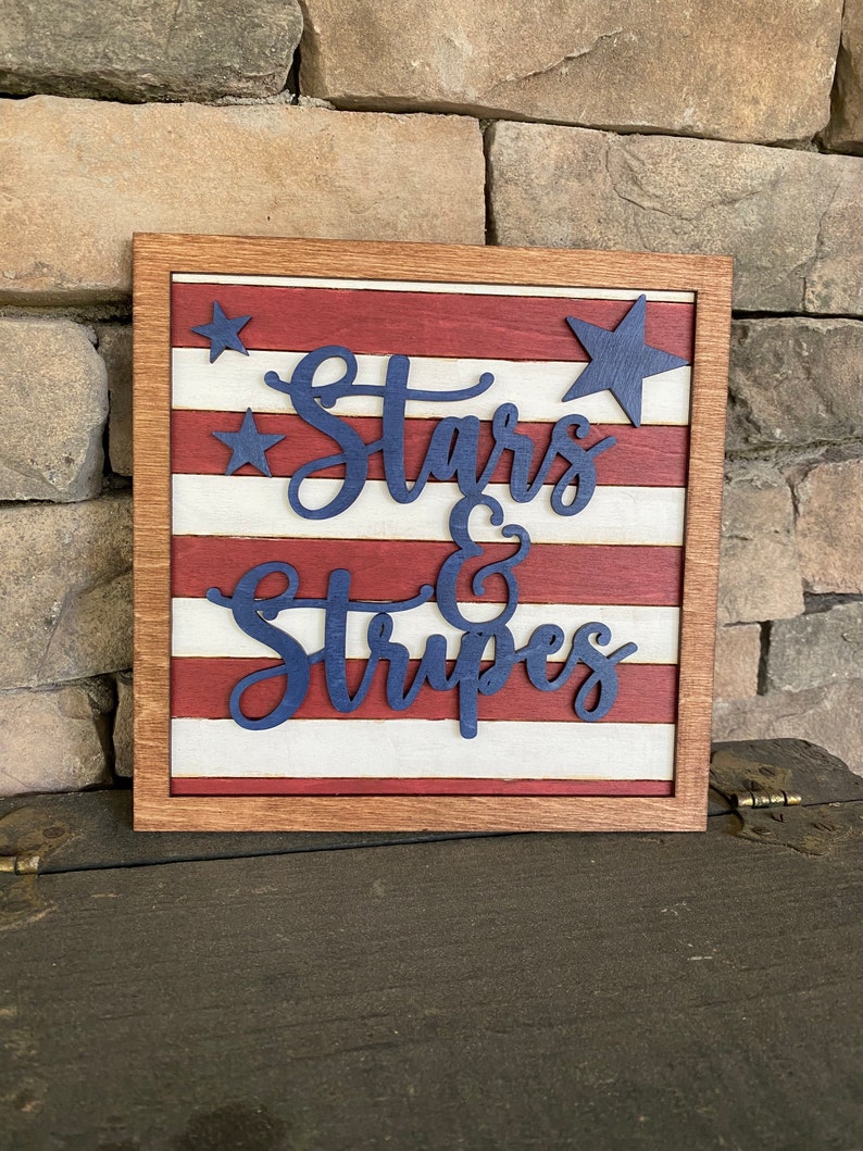 Puede incluir: Un letrero de madera con un dise&ntilde;o de bandera estadounidense roja, blanca y azul. El letrero dice "Stars & Stripes" en letras azules.