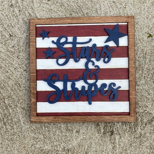 Puede incluir: Un letrero de madera con un dise&ntilde;o de bandera estadounidense roja, blanca y azul. El letrero dice "Stars & Stripes" en letras azules.