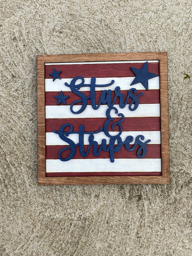Puede incluir: Un letrero de madera con un dise&ntilde;o de bandera estadounidense roja, blanca y azul. El letrero dice "Stars & Stripes" en letras azules.