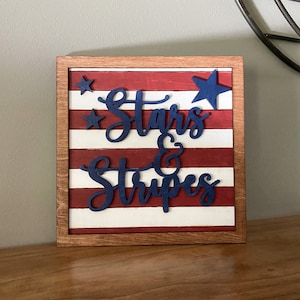 Puede incluir: Un letrero de madera con un dise&ntilde;o de bandera estadounidense roja, blanca y azul. El letrero dice "Stars & Stripes" en letras azules.