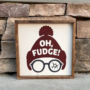 May include: A wooden sign with a white background and a red and white illustration of a person wearing a hat and glasses. The text "OH, FUDGE!" is written in white on the hat.