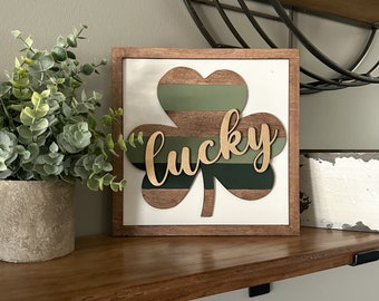 May include: Wooden sign with a shamrock design. The shamrock has green and brown stripes, with the word "lucky" in a cursive font. The sign is framed in a brown wooden border and sits on a wooden shelf.