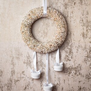 May include: A white dried flower wreath with four white boxes hanging from it. The wreath is hanging on a beige textured wall.
