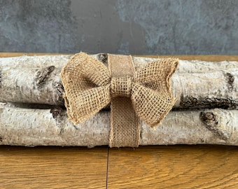 May include: Three birch logs tied together with a brown burlap ribbon and bow. The logs are stacked on top of each other and the ribbon is tied around the middle of the stack.