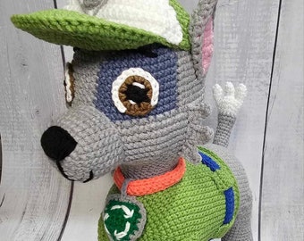 Op de afbeelding: Een gehaakte knuffelhond die een groen shirt draagt met een recycle-symbool, een groen-witte baseballpet en een oranje halsband. De hond heeft grijs haar, witte poten en een roze oor.