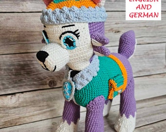 Op de afbeelding: Gehaakt knuffeldier dat lijkt op een hond met paarse vacht, witte poten en een groen-oranje vest. Het speelgoed draagt ​​een blauw-witte hoed met een roze oor.