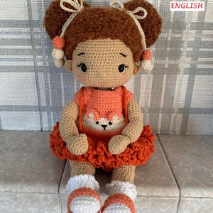 Op de afbeelding: Een handgemaakte gehaakte pop met bruin haar in twee knotjes, versierd met crèmekleurige strikken en perzik-kleurige kralen. De pop draagt een oranje jurk met een wit vossenontwerp en een bijpassende rok. Witte en oranje sandalen maken de outfit compleet.