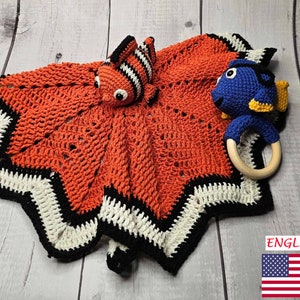May include: A crocheted baby blanket with a star-shaped border in orange, white, and black. The blanket features two crocheted stuffed toys: a clownfish and a blue fish with a wooden ring.