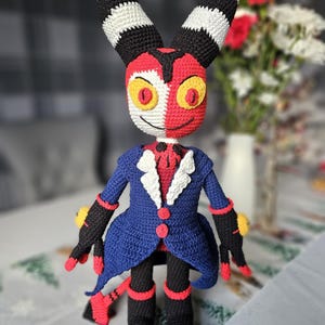 Puede incluir: Un juguete de peluche tejido a crochet de un personaje con orejas rayadas en blanco y negro, una cara roja y blanca y un traje azul. El juguete tiene ojos amarillos y una sonrisa traviesa.